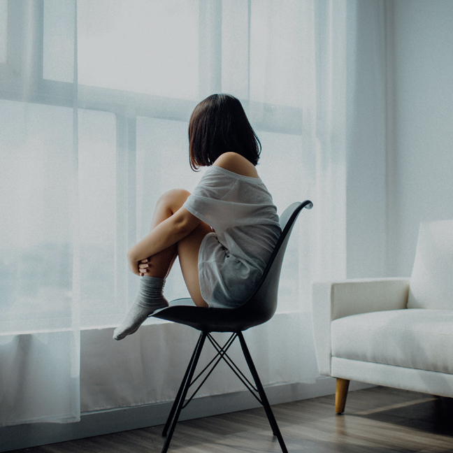 A child sitting alone on a chair by a window, hugging their knees.